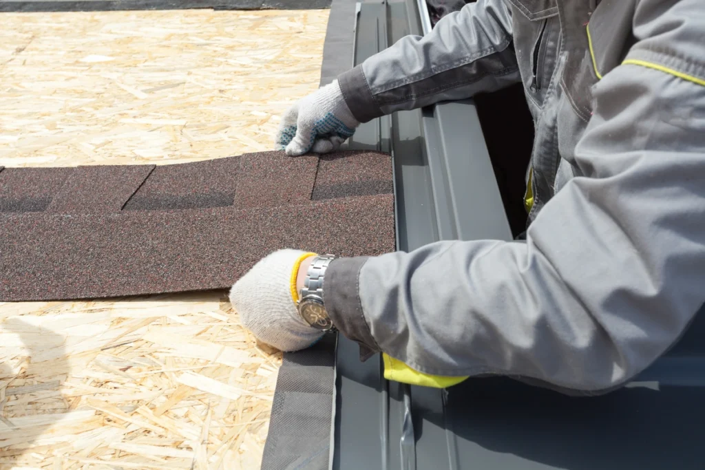 A construction worker, carefully aligning brown asphalt shingles at the roof edge, dons gloves and a gray work jacket. As he meticulously finishes the wooden deck's installation, homeowners might wonder: do you need a permit for a roof replacement?