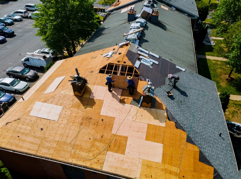 Aerial view of workers replacing a roof on a large building, with part stripped to bare wood and new shingles installed—raising the question: how long does a 30 year roof really last? Construction materials and vehicles are visible nearby.