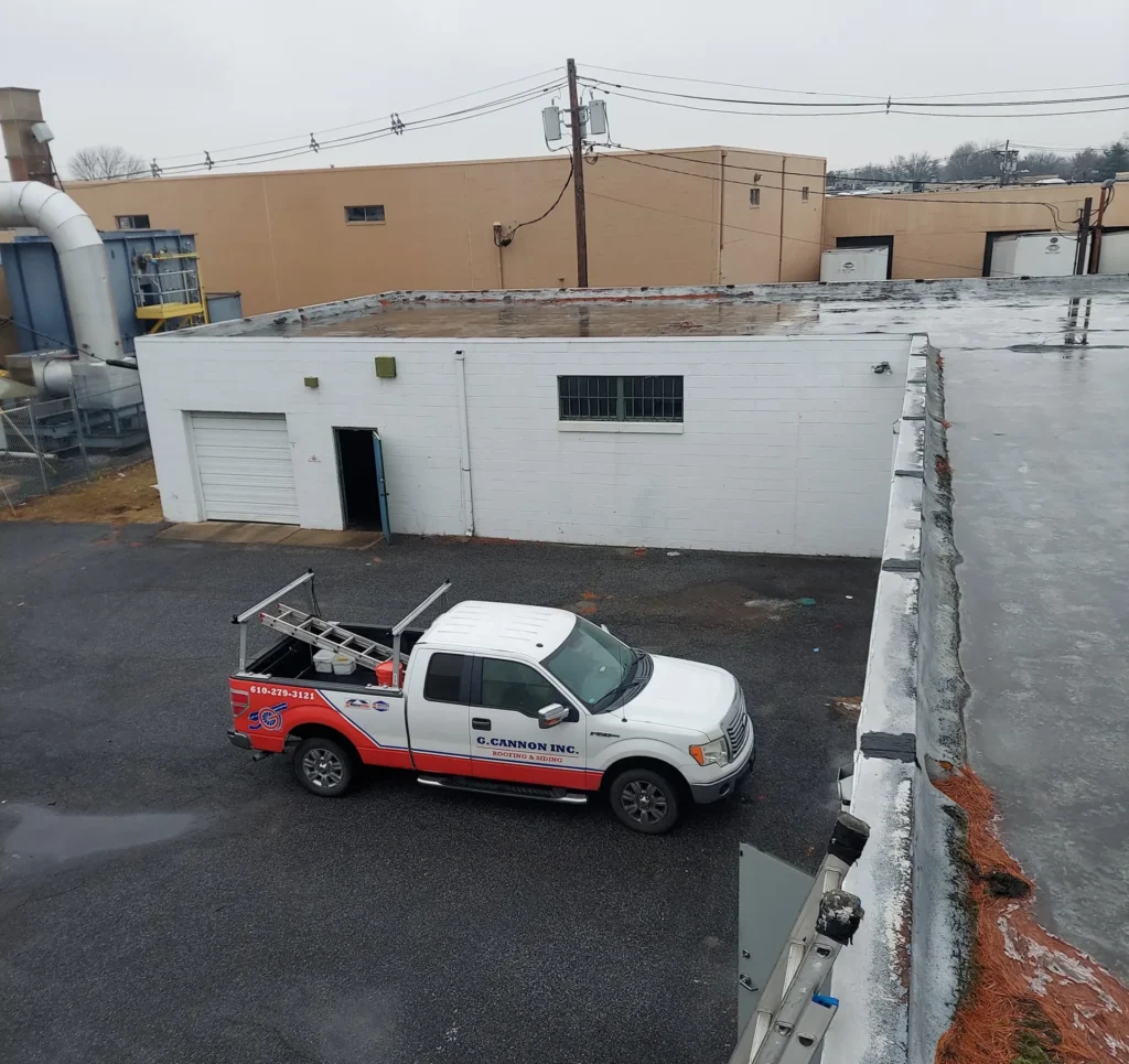 A white and red work truck is parked in an industrial lot next to a white building. The scene is overcast, with damp pavement suggesting recent rain. A ladder is mounted on the truck's side, and buildings and utility poles are visible in the background.