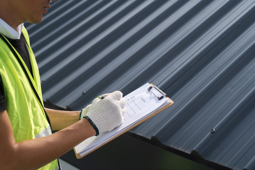 A person wearing a safety vest and white gloves holds a clipboard and pen, inspecting or taking notes next to a dark-colored metal roof