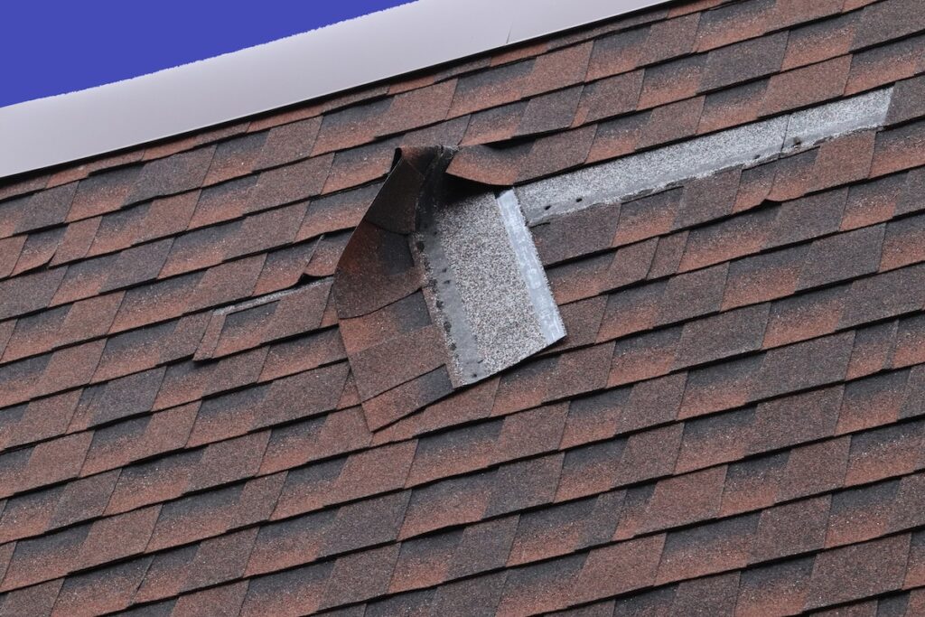 A close-up of a house roof with brown asphalt shingles, showing visibly damaged and curling shingles near a roof seam or ridge