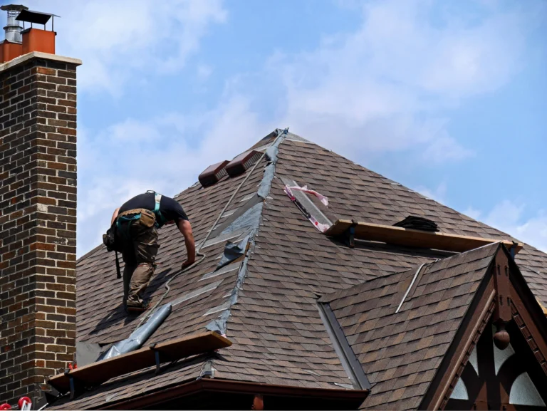 A worker wearing a safety harness is repairing a steep, shingled roof on a brick house, addressing various roof damage types. The sky is partly cloudy, and there are wooden planks and tools scattered around. A brick chimney is visible on the left side.