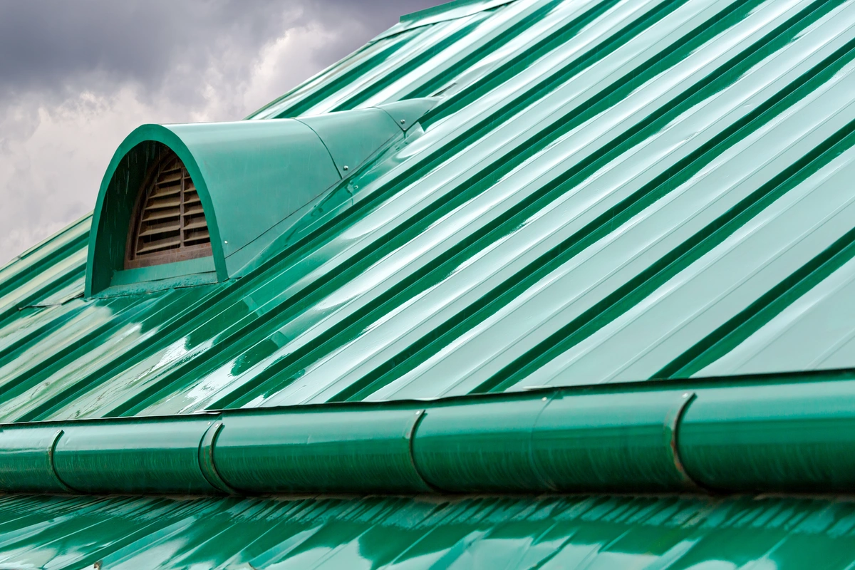 A close-up view of a green metal roof with a curved vent structure against a cloudy sky background. The roof panels, adorned in one of many striking metal roof colors, are shiny and have a ridged texture.
