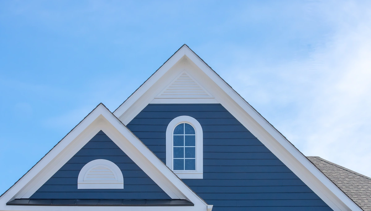 The image showcases a light blue sky framing the gabled roof of a blue house with white trim, echoing CertainTeed siding colors. Two prominent gables stand out: one with a semi-circular window and another featuring a tall, narrow arched window.