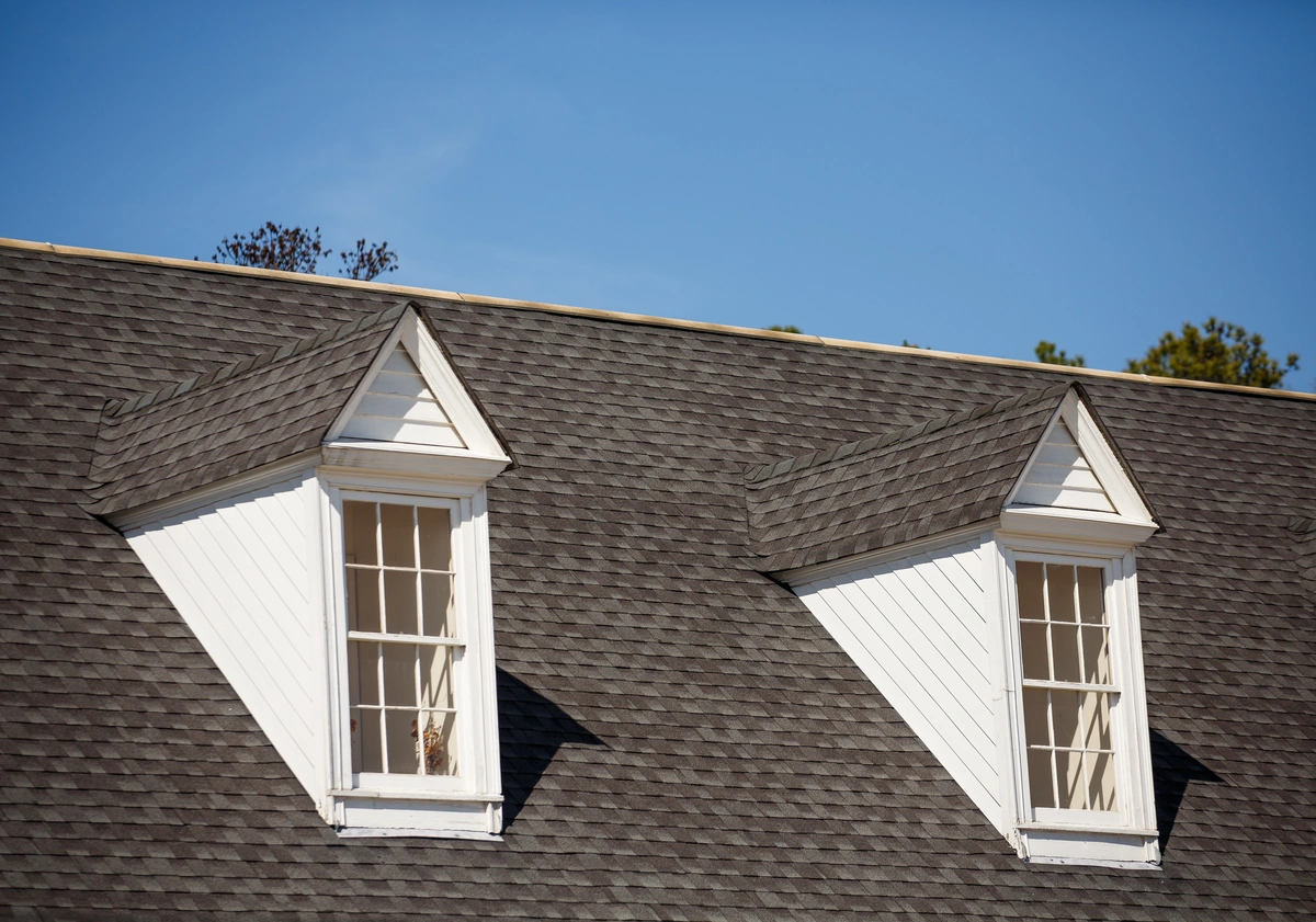 A roof with two white dormer windows against a clear blue sky showcases the best roof shingles. The dark gray shingles are neatly arranged, and the white trim around the windows contrasts with the roof. Trees are partially visible in the background.