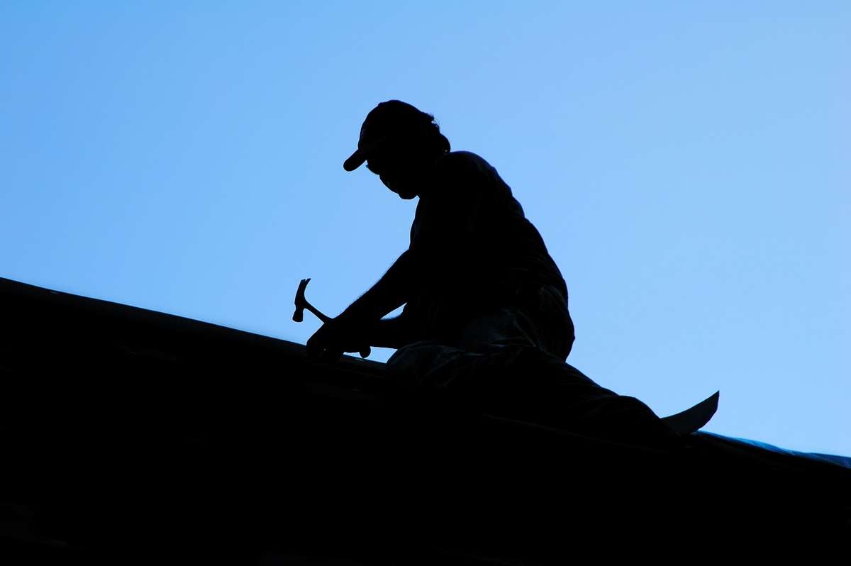 Silhouette of a person wearing a cap and expertly wielding a hammer on a rooftop against a clear blue sky, embodying the skills needed when considering how to choose a roofer.