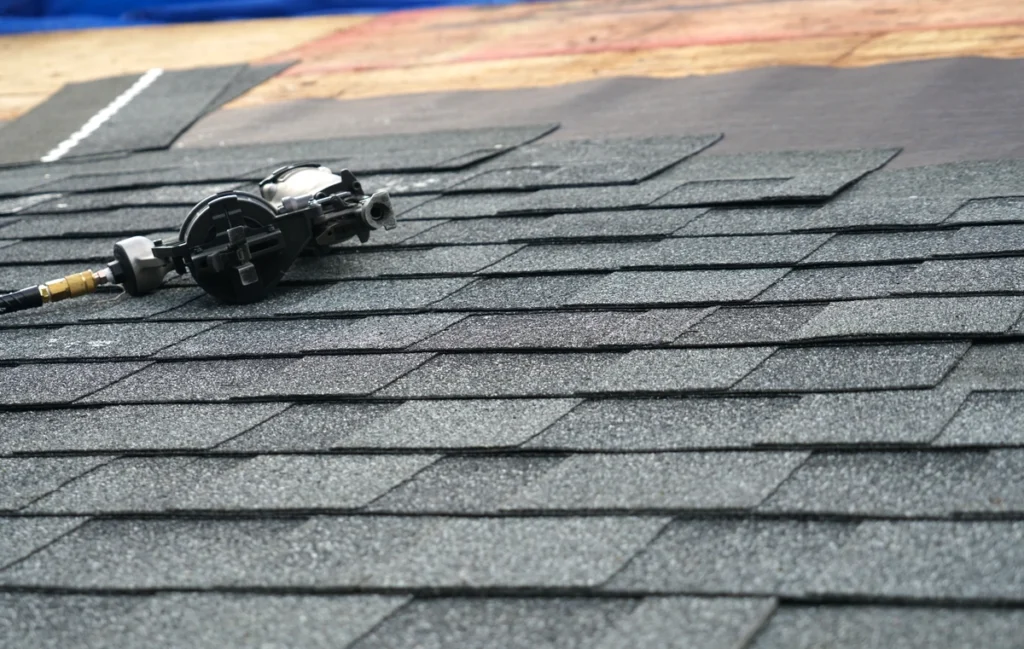 Close-up of a partially shingled roof with a pneumatic nail gun resting on the asphalt shingles, illustrating how to measure a roof for shingles. The background reveals the tar paper underlayment and wooden decking, essential components in roofing projects.