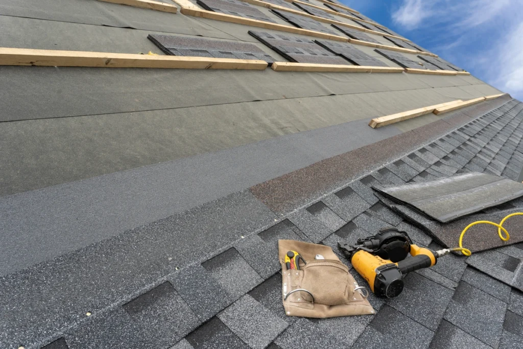 A roof in the process of being shingled shows layers of black and gray shingles, illustrating techniques on how to measure a roof for shingles. Wooden planks and a nail gun with nails are scattered about, while the clear blue sky stretches in the background.