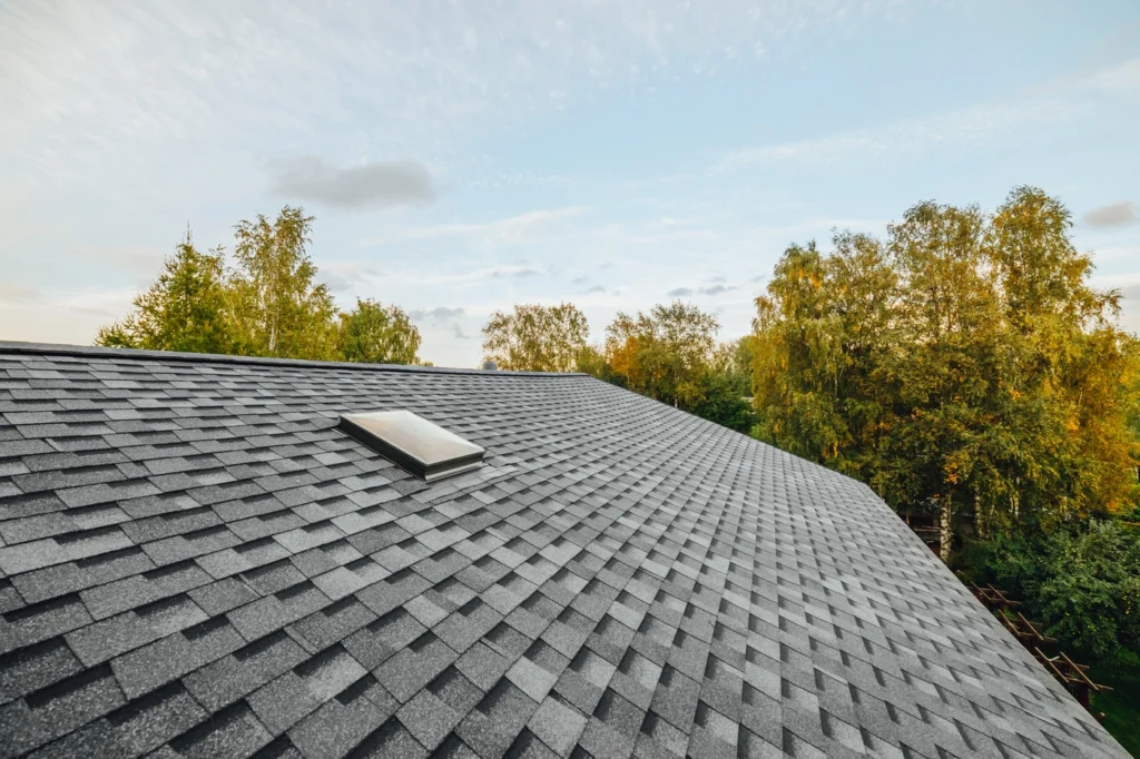 A rooftop with gray asphalt shingles, expertly installed by Conshohocken Roofing, features a small skylight. The background showcases lush green trees set against a blue sky dotted with scattered clouds.