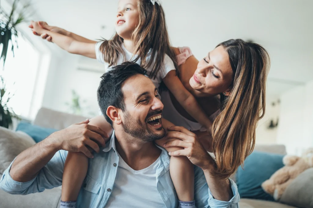 A joyful family moment unfolds in a cozy, well-lit living room: a man smiles as a little girl sits on his shoulders, playfully reaching out. Beside them, a woman stands smiling warmly, perhaps enjoying the peace of mind that comes with her reliable Phoenixville roofing keeping their home snug and secure.