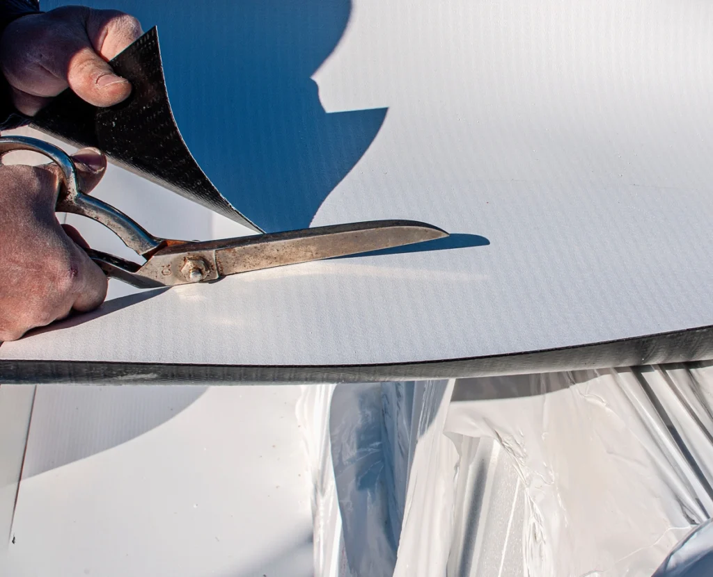 Close-up of a person cutting a large sheet of white material with metal scissors, akin to the precision needed for crafting a TPO roof. The sheet rests on a surface covered with a protective plastic layer. The scene is well-lit, highlighting the texture of the material.