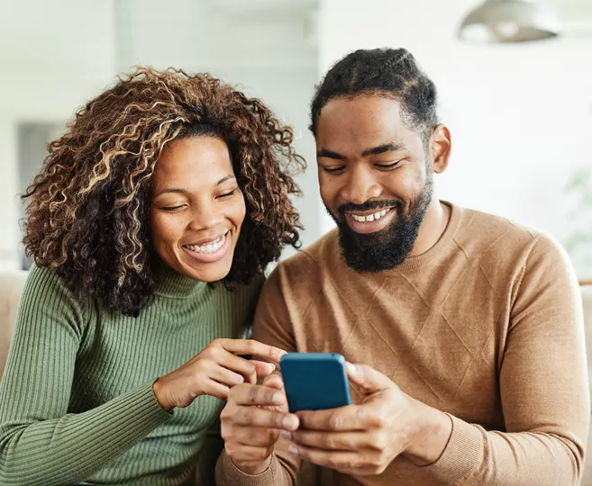 A smiling couple sits closely on a couch, looking at a smartphone together. The woman is wearing a green sweater, and the man is in a brown sweater. Perhaps they're exploring reviews for Blue Bell Roofing, given their engaged and happy expressions while sharing this activity.