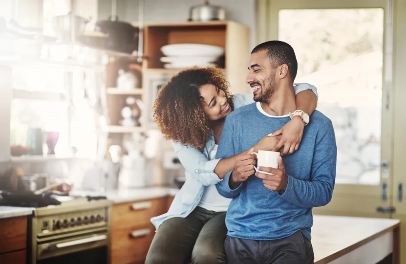 In a cozy kitchen meeting, a couple savors a tender moment. The woman sits on the counter, hugging the man from behind as he holds a mug. Their smiles convey warmth and happiness, with kitchen appliances and glimpses of the newly finished roofing visible in the background.