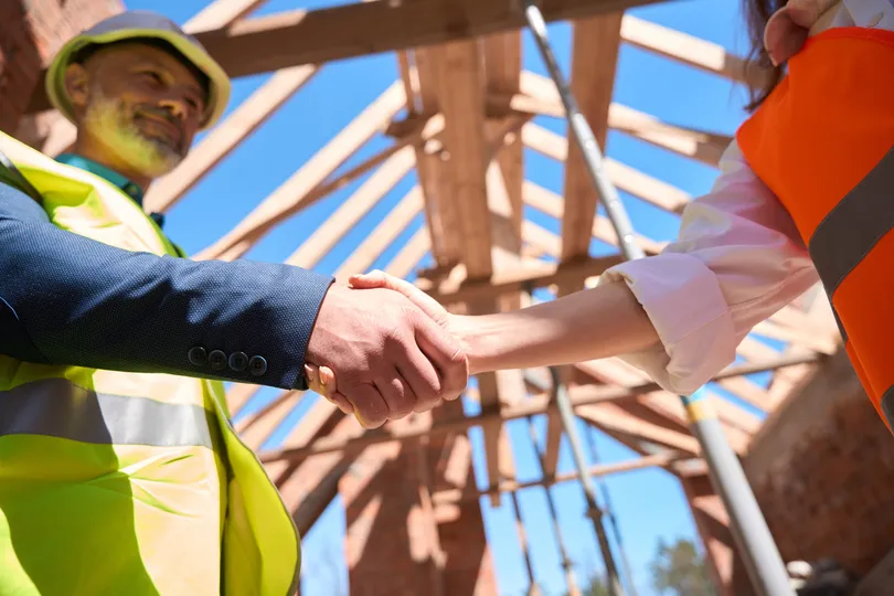 Two construction workers, one wearing a yellow vest and helmet and the other in an orange vest, are shaking hands at a construction site in Conshohocken, PA. Behind them, a wooden roof frame stands under a clear blue sky, promising quality commercial roofing ahead.