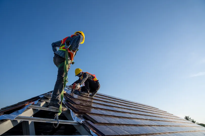 Two workers in safety gear and helmets install solar panels on a sloped roof under a clear blue sky, showcasing Collegeville Commercial Roofing's commitment to sustainable energy. They stand on the panels, with one kneeling and adjusting an attachment, embodying eco-friendly solutions.
