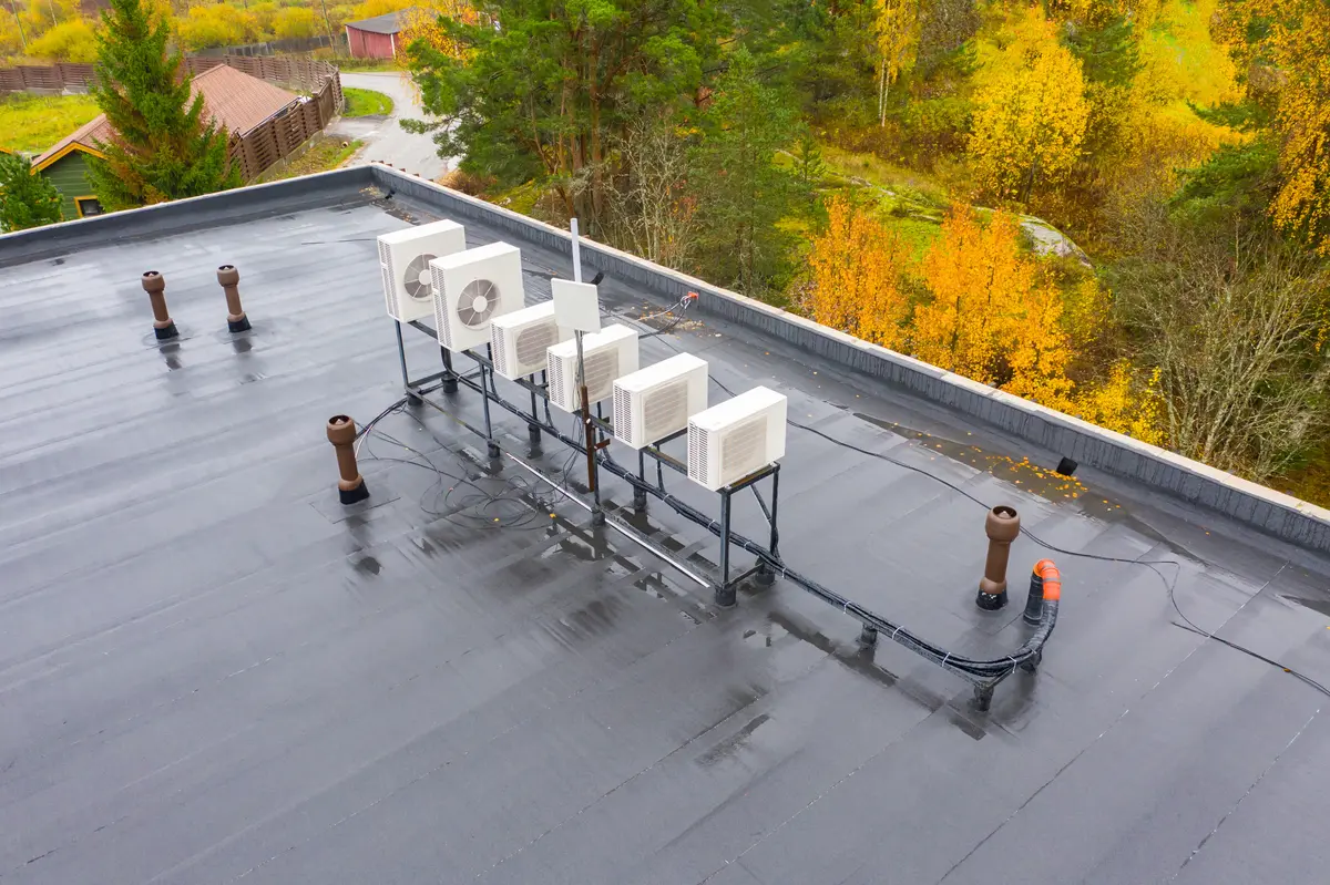 The rooftop, showcasing one of the many types of commercial roofing, features six HVAC units on metal supports atop a flat, black surface. Surrounded by autumn trees with yellow and green leaves, the scene is set under overcast skies with a wet roof. A road and wooden building complete the background.