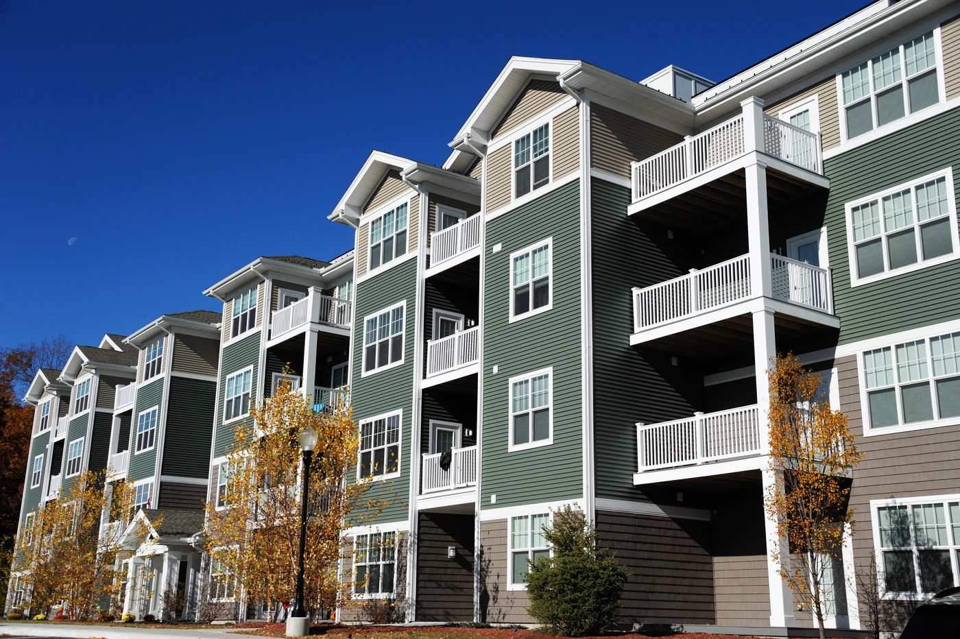 Four-story apartment building with green and beige siding, featuring multiple balconies. The architecture includes white trim and windows. Trees with autumn foliage are in the foreground under a clear blue sky.