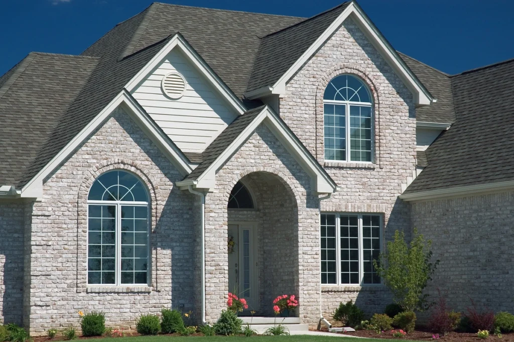 Beautiful suburban home with a stone and siding exterior, featuring arched windows and a shingled roof by Havertown Roofing. A well-manicured lawn and small flower garden enhance its curb appeal under a clear blue sky.