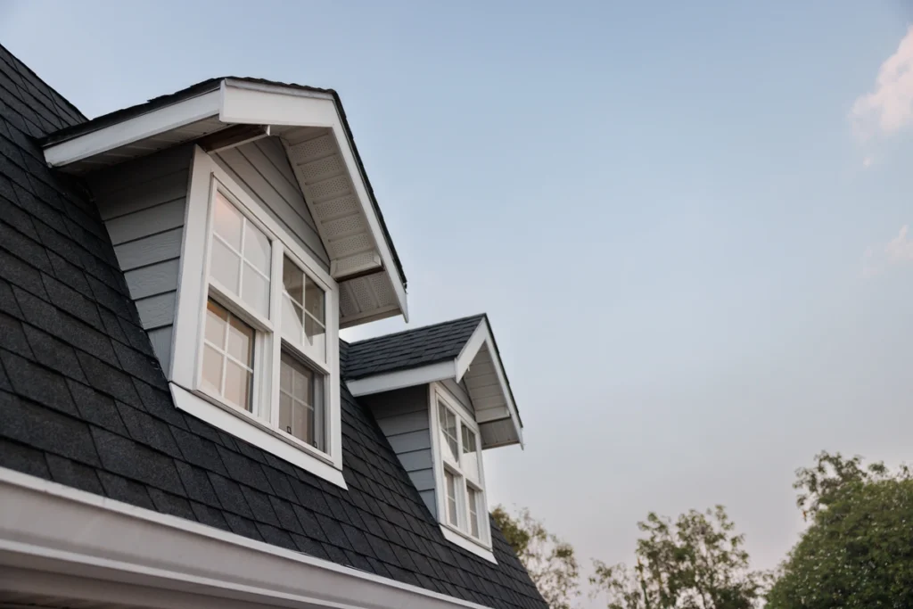 A close-up view of a house's roof, showcasing Berwyn Roofing's craftsmanship, features two dormer windows with white trim set against a clear blue sky. The roof is covered with dark shingles, and trees are visible in the background.