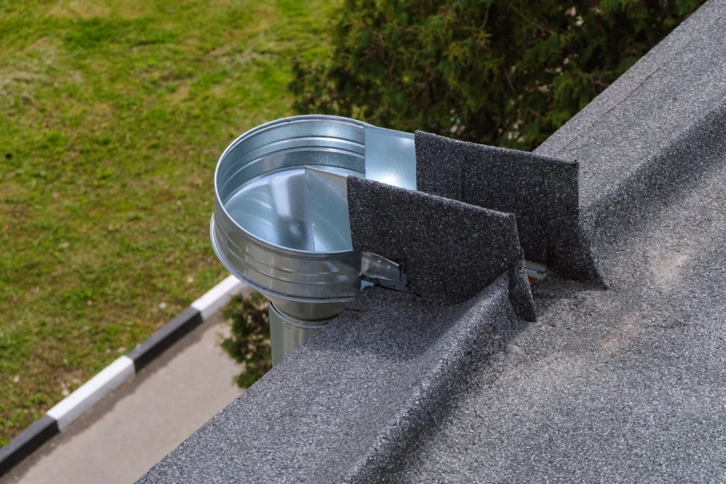 A metal rain gutter on a flat rooftop with asphalt shingles, directing water into a circular downspout. The surrounding area includes grass, a tree, and a partially visible road with a curb.