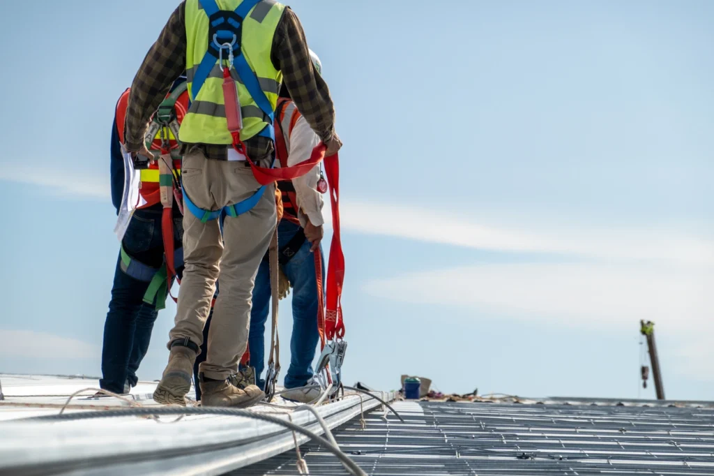 Workers in safety gear and harnesses stand on a rooftop. They appear to be installing or inspecting metal roofing