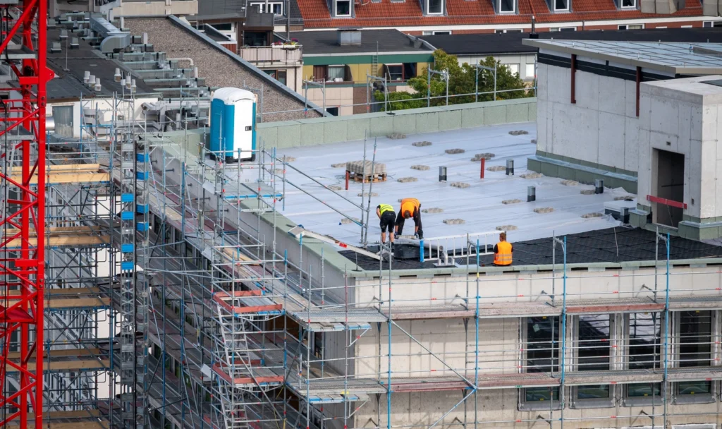 Construction workers on a rooftop perform tasks on a building under construction. Scaffolding surrounds the structure, and a red crane is visible on the left