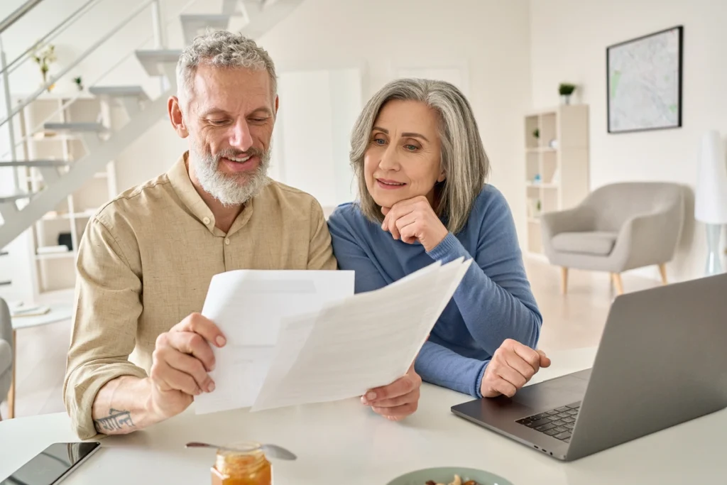 An older couple sits at a table, reviewing documents together, possibly for a roofing service in Hatboro. The man holds the papers while the woman looks on thoughtfully. A laptop is open beside them, and there's a jar of jam and some nuts nearby. The room is bright and modern.