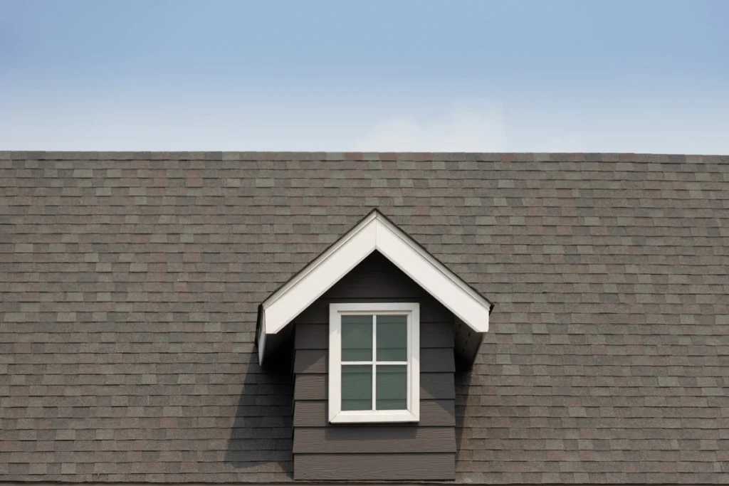 A gabled dormer window with white trim elegantly protrudes from a professionally installed Royersford roofing job, its grey shingle roof contrasting beautifully against the clear blue sky.