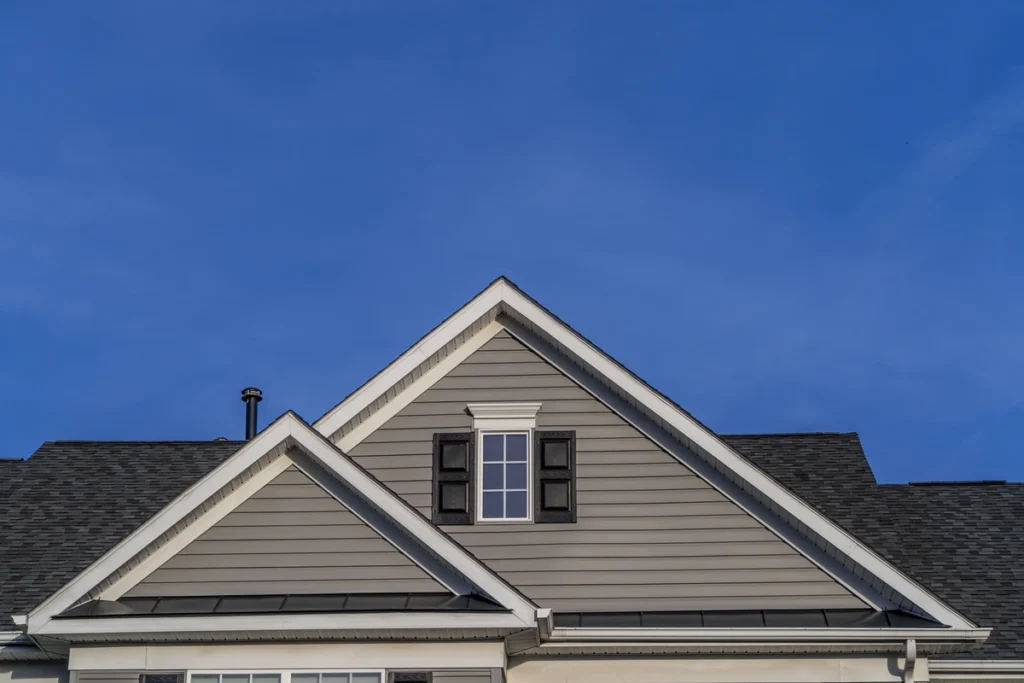 A close-up of the roof of a house with gray siding and black shutters against a clear blue sky showcases the expert craftsmanship of Roofing Team Broomall. The design features a gable roof with white trim, highlighting their attention to detail.