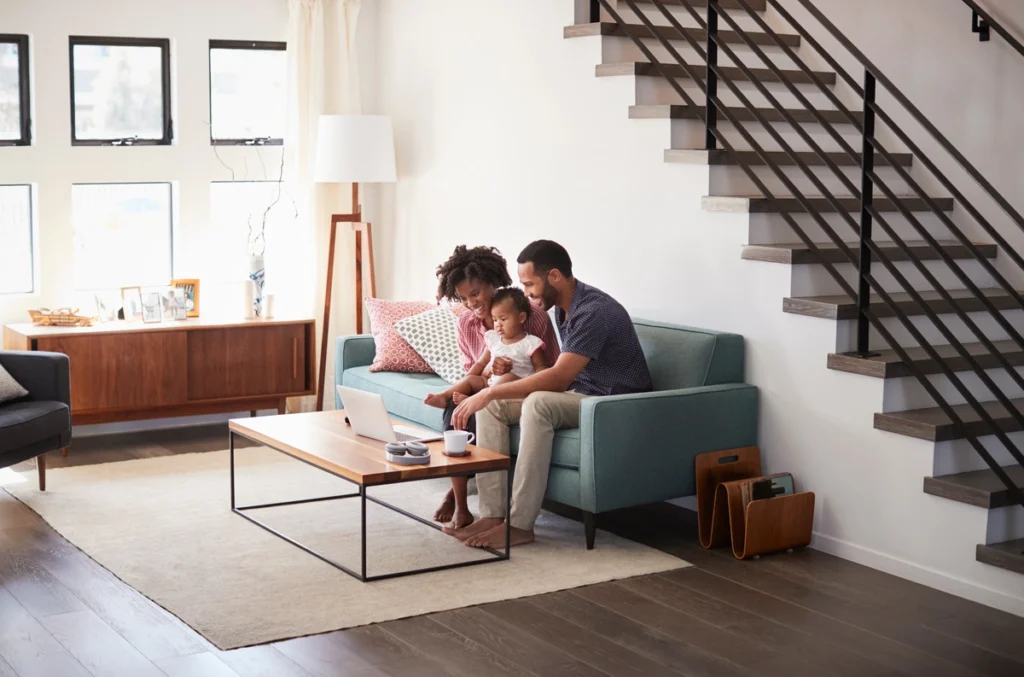 A family of three sits on a teal sofa in a modern living room by Eagleville Roofing. The parents engage with their child, who is holding a toy. The room features a minimalist design with a wooden coffee table, sideboard, and open staircase.