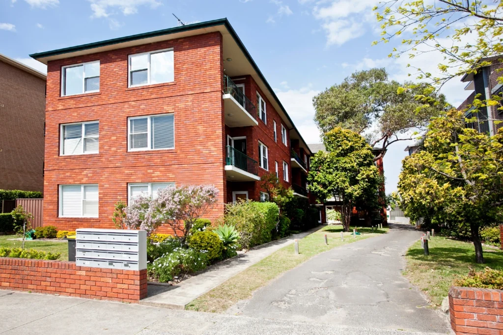 A three-story red brick apartment building with balconies, surrounded by greenery and trees. A narrow driveway runs beside it, leading to the rear.