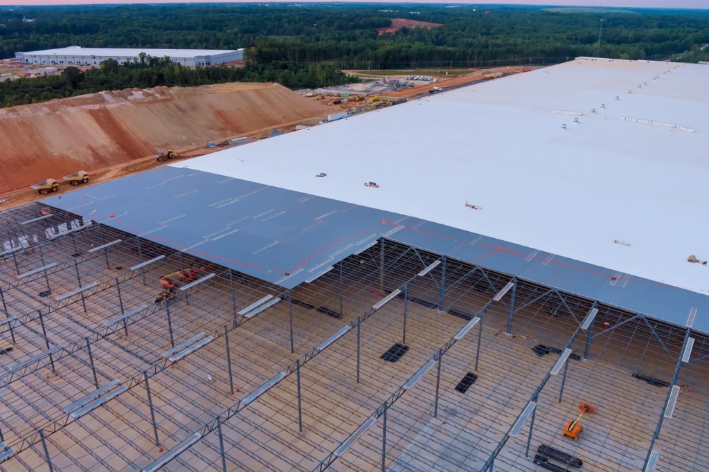 Aerial view of a large industrial building under construction. The structure features a vast roof, partially completed, with visible steel framework