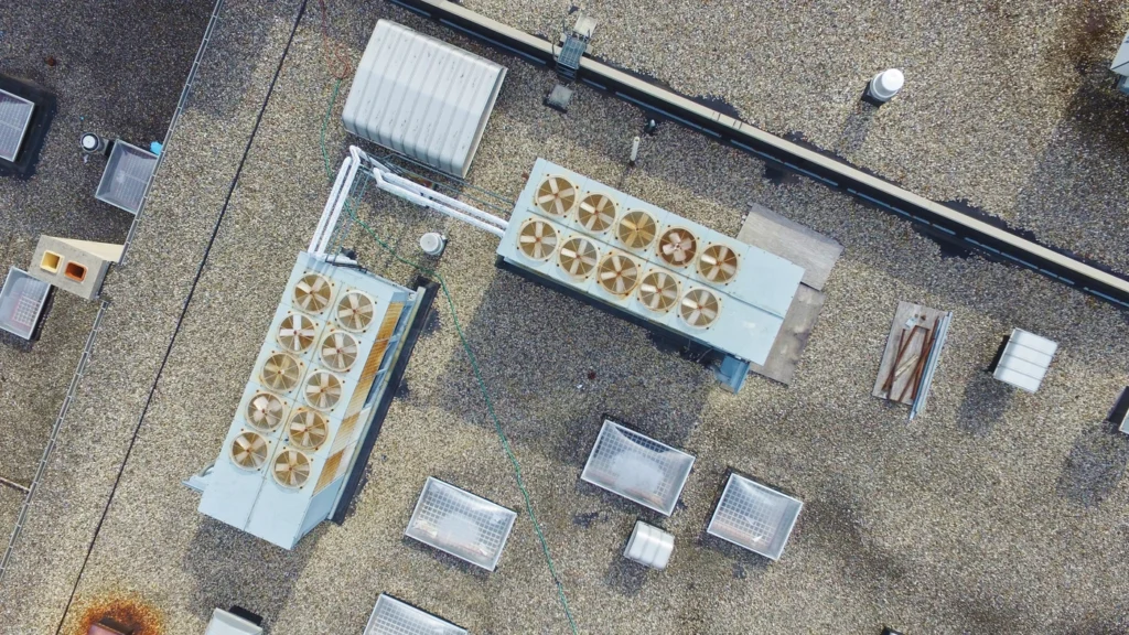 Aerial view of a flat, pebble-covered rooftop with two large HVAC units featuring multiple fans. Several rectangular skylights and utility structures are also present, along with various pipes and vents.
