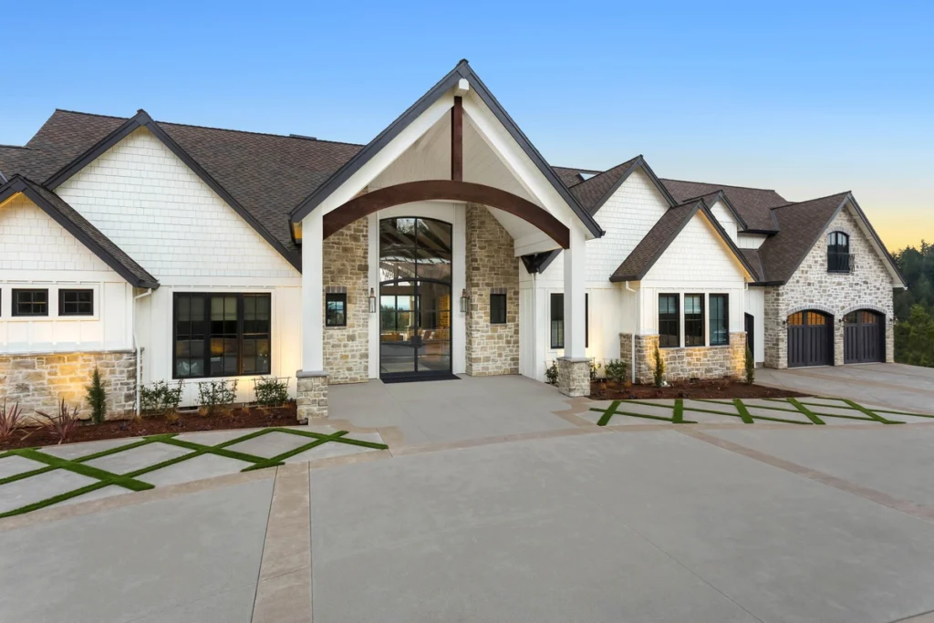 A large modern house with a stone and white exterior under a blue sky showcases expert roofing services Horsham trusts. It features multiple gabled roofs, large windows, and a wide driveway leading to an arched entrance. The surrounding area is beautifully landscaped with grass and plants.