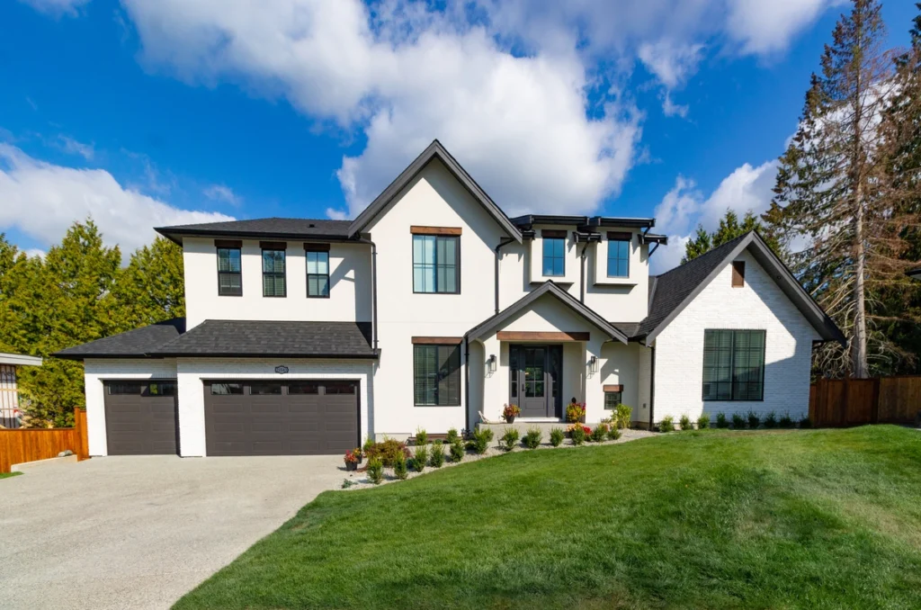 A modern two-story house in Ambler, PA, boasts white walls and a dark roof. It features a two-car garage and a well-maintained lawn. Tall trees stand gracefully in the background under a partly cloudy sky.