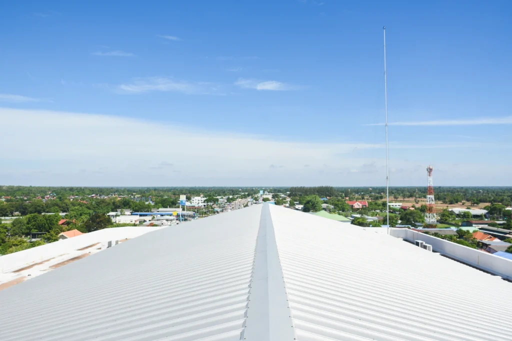 View from a rooftop with a white metal surface, leading to a vast landscape with greenery, houses, and a distant horizon. The sky is clear and blue, featuring a tall radio tower on the right.