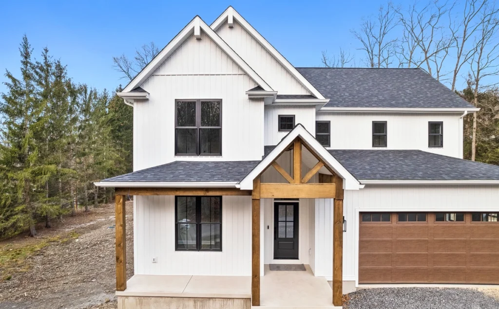 A modern two-story house in Ambler, PA, boasts a sleek white exterior and a stylish dark roof. It features a covered porch with wooden beams and a double garage with wooden doors. Nestled among trees, this home offers serene charm with its gravel driveway.