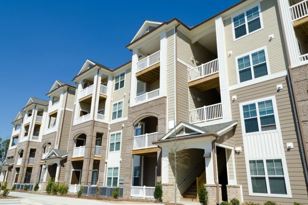 A row of modern apartment buildings under a clear blue sky. The buildings feature multiple stories, balconies, and a combination of brick and siding exteriors