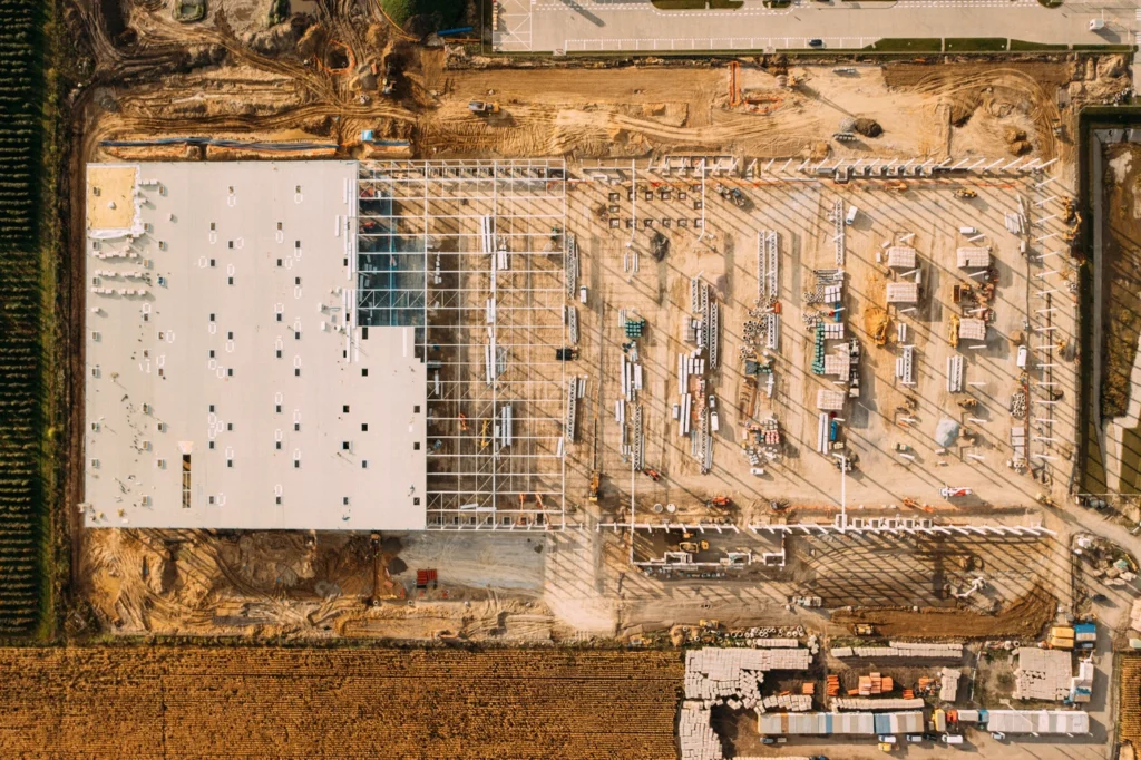 Aerial view of a large construction site with a partially completed building. The structure has a white roof, exposed steel framework, and scattered construction materials