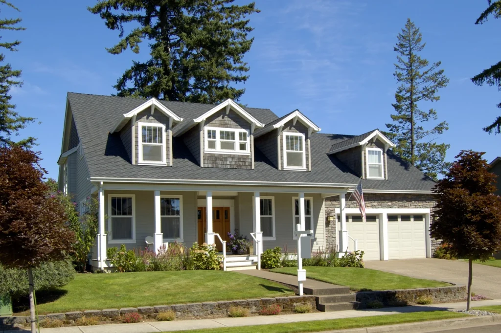 A charming two-story house with a gray exterior, featuring expert Doylestown roofing, a large front porch, and manicured landscaping. There are three dormer windows on the roof, an attached two-car garage, and a U.S. flag next to the entrance. Tall trees surround the property.