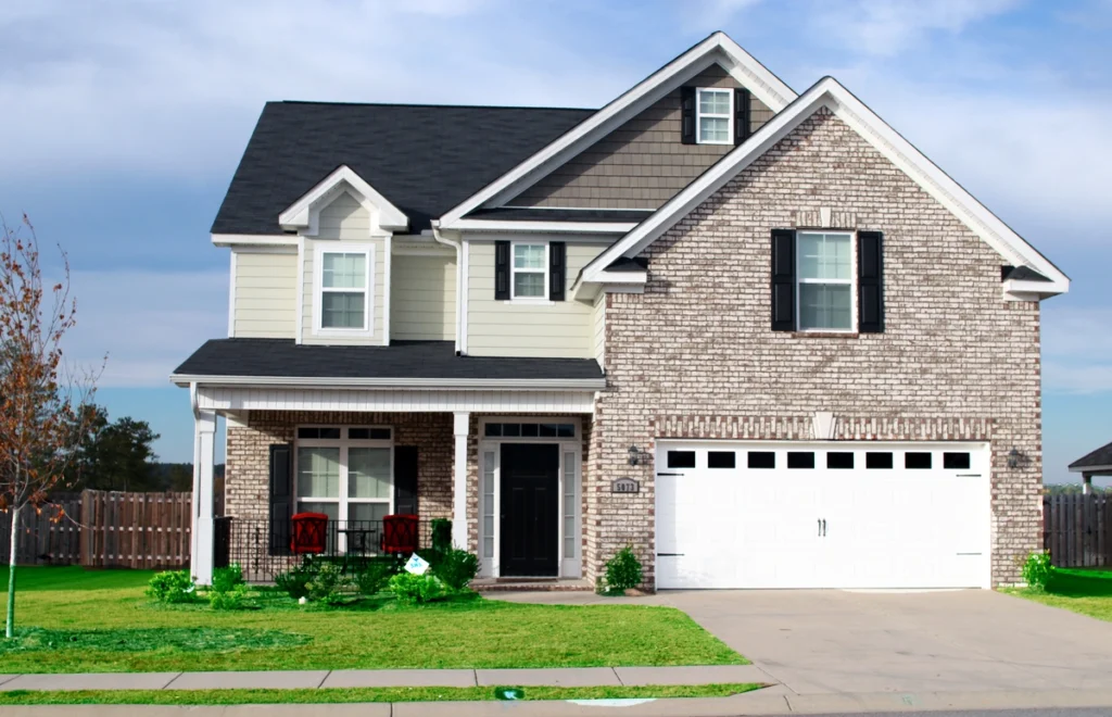 A charming two-story suburban house features a brick and siding exterior, complemented by a black front door and white garage. With king of Prussia roofing adding durability, the home boasts a cozy front porch with two chairs, a lush lawn, and a wooden fence under a partly cloudy sky.