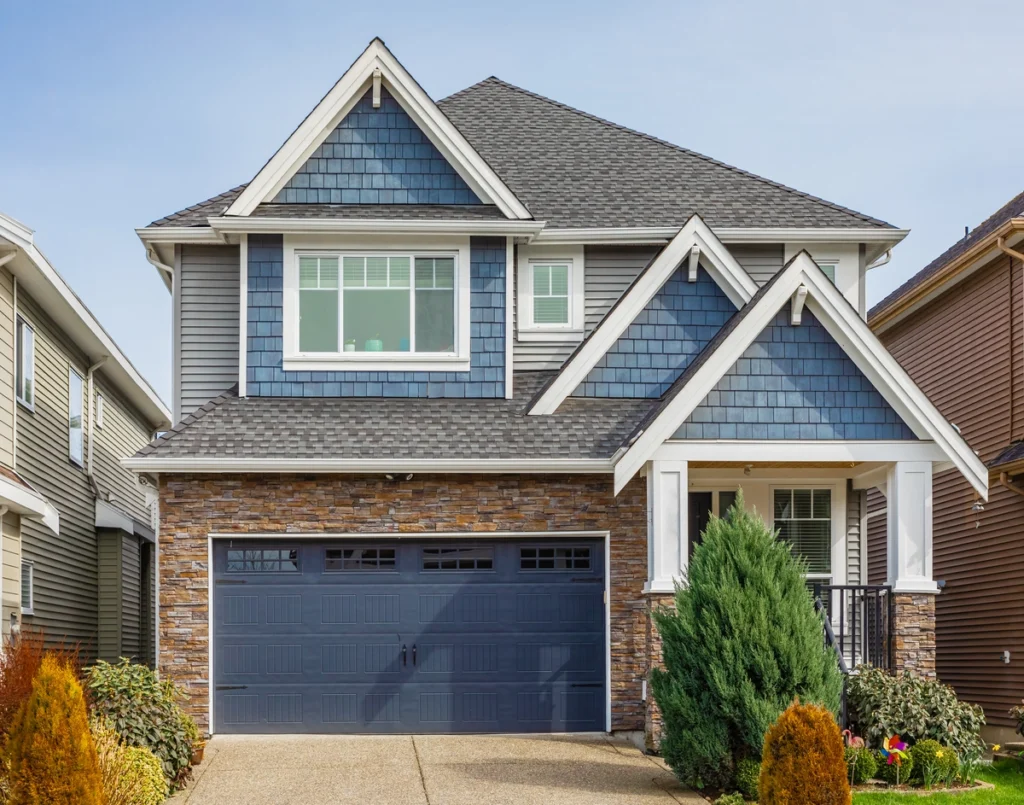 A modern two-story house with blue and gray siding, a double garage, and a stone facade showcases excellent Collegeville roofing. A well-maintained front yard with shrubs and a small porch is visible under a clear sky.