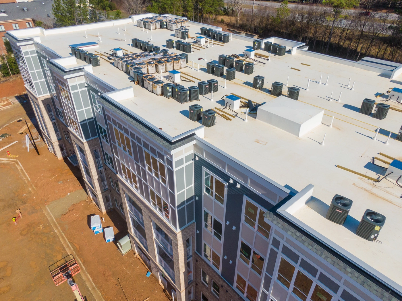 Aerial view of a large, white-roofed building under construction. The rooftop is equipped with multiple air conditioning units and other installations. Surrounding the building is undeveloped land with some trees in the background.