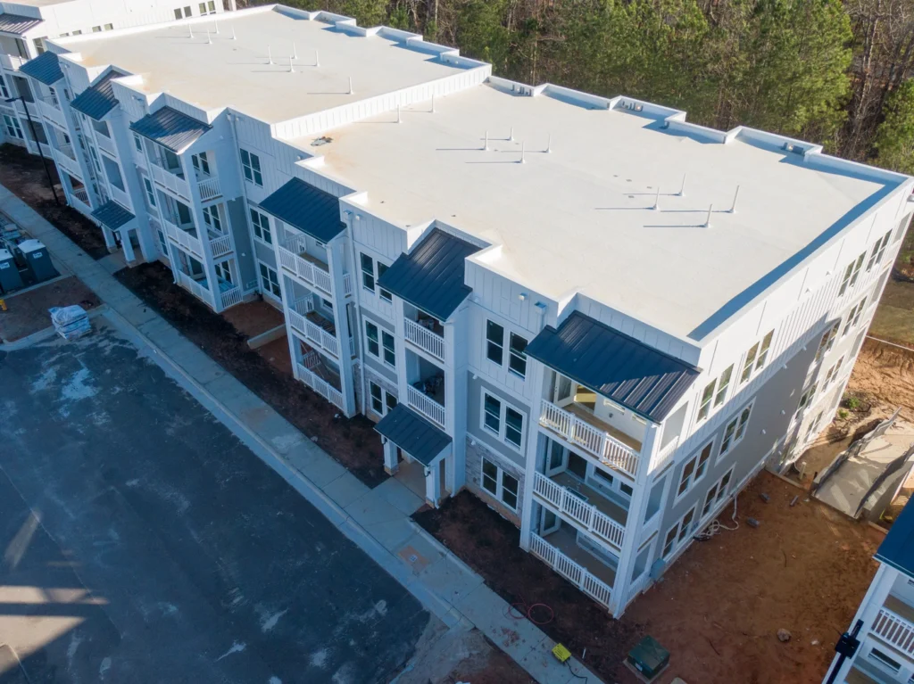 Aerial view of a newly constructed apartment complex with multiple three-story buildings featuring white exteriors and dark roofs. The area around the buildings includes partially developed landscaping and a parking lot.