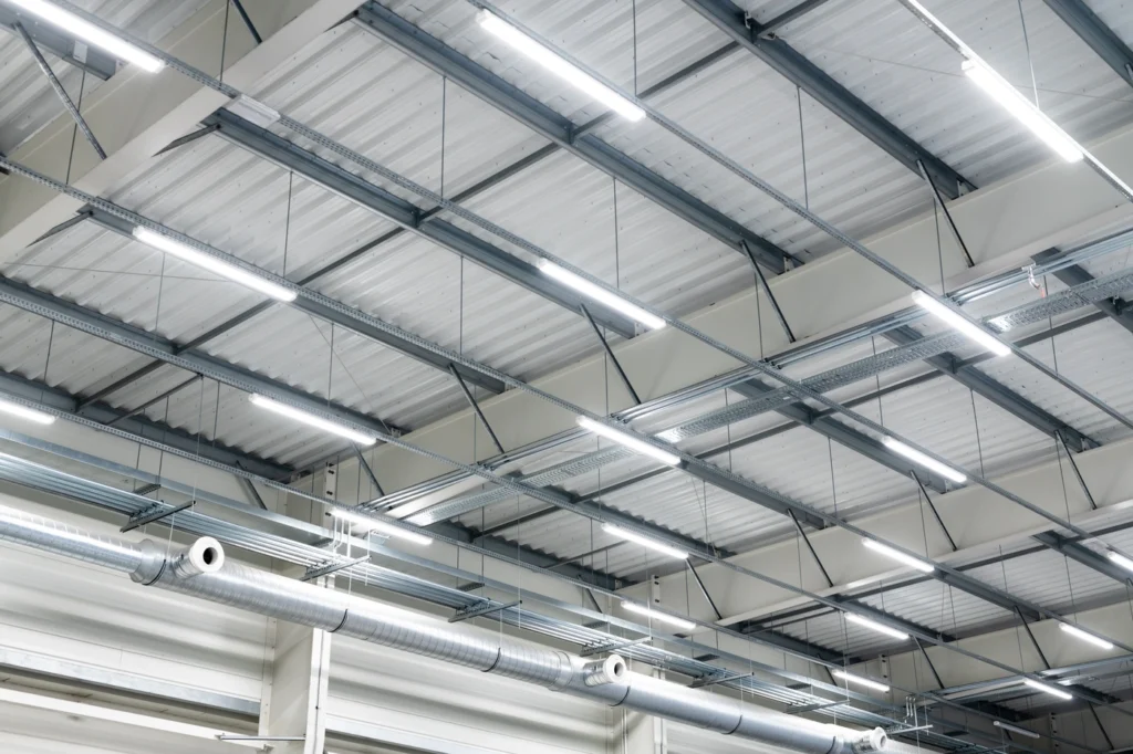 View of a large warehouse ceiling with a grid of fluorescent lights, metal beams, and ventilation ducts. The ceiling panels are white, contributing to a clean, industrial look.