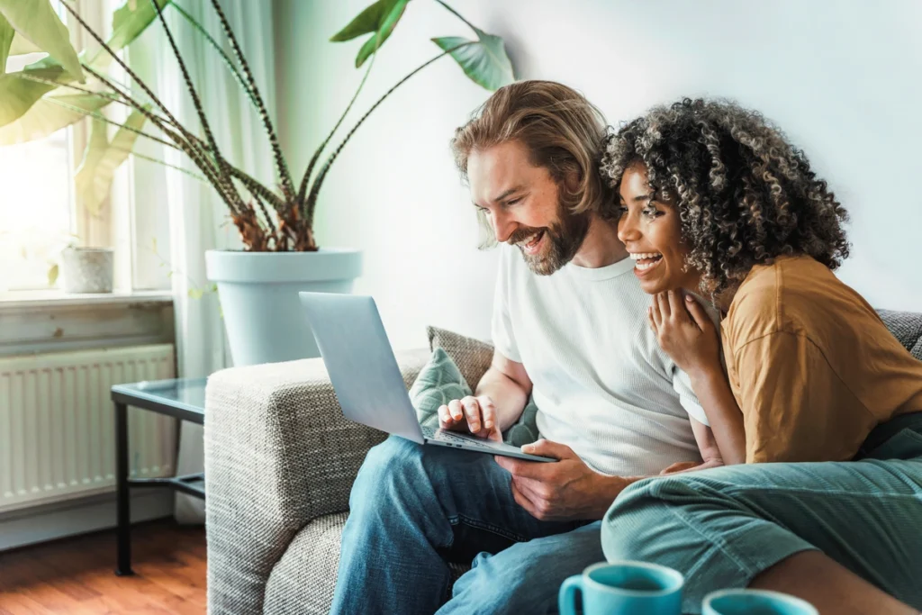 A smiling couple sits on a couch in bright living room, exploring Collegeville roofing options on a laptop. A large potted plant graces the background, while two mugs rest on the table in the foreground.