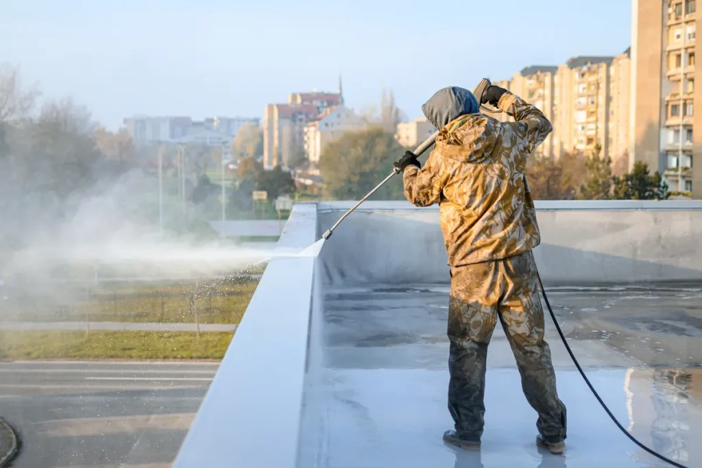 A person in camouflage gear pressure washes a flat rooftop in an urban area. Tall buildings and trees are visible in the background under a clear sky.