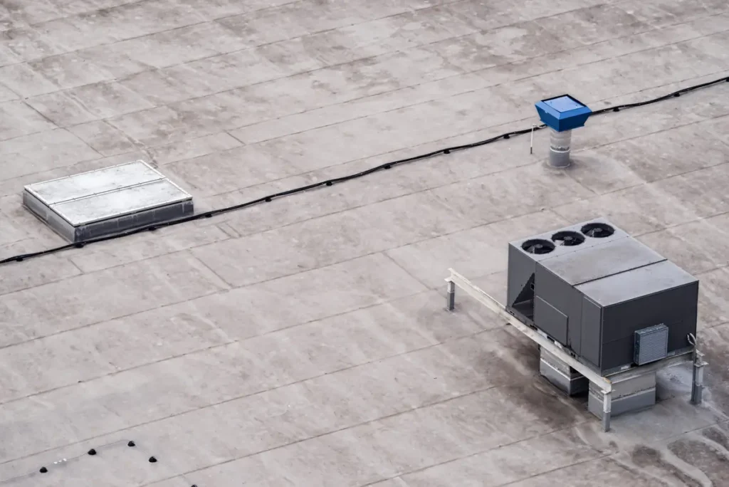 Aerial view of a flat rooftop with industrial equipment, including an HVAC unit and a small blue ventilation structure. The surface is mostly light gray, and there are linear pathways suggesting utility conduits.