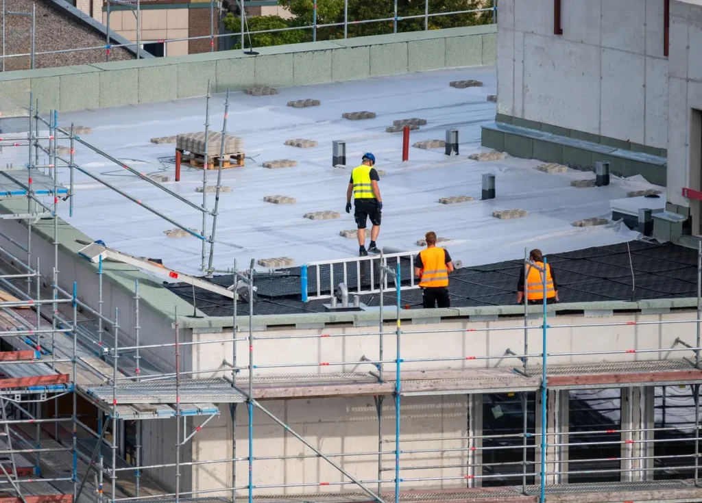 Three construction workers in high-visibility vests work on a building roof. One is carrying a ladder, another is walking, and the third is observing. Scaffolding surrounds the building, and the roof is partially covered with protective material.