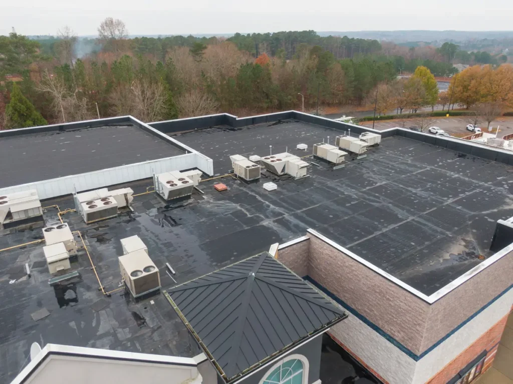 Aerial view of a large flat rooftop with multiple HVAC units. The building is surrounded by trees with autumn foliage and a road with parked cars. The sky is overcast.